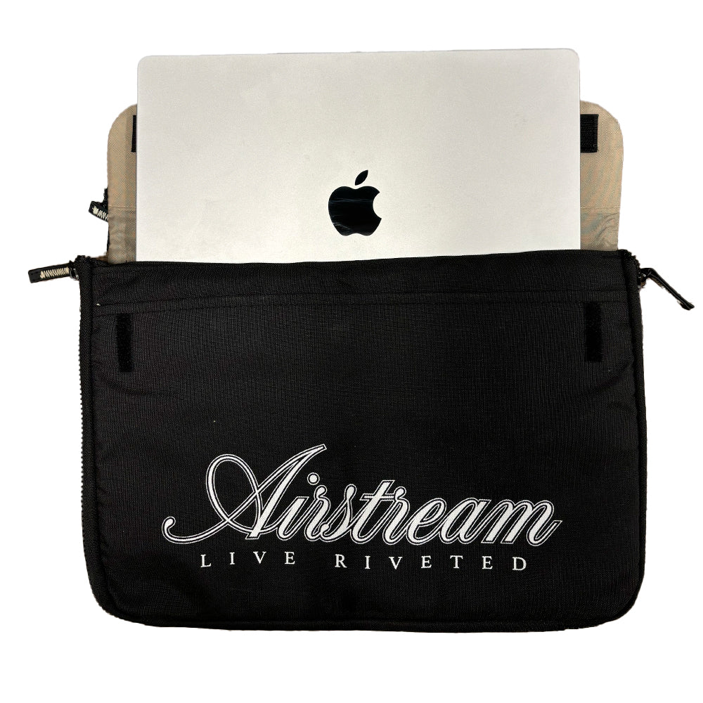 A silver laptop with the Apple logo is partially inside a black zippered pouch that has “Airstream LIVE RIVETED” written on it in white script. The Airstream Signature Series Laptop Commuter Sleeve features a padded laptop compartment, offering both style and protection for your device.