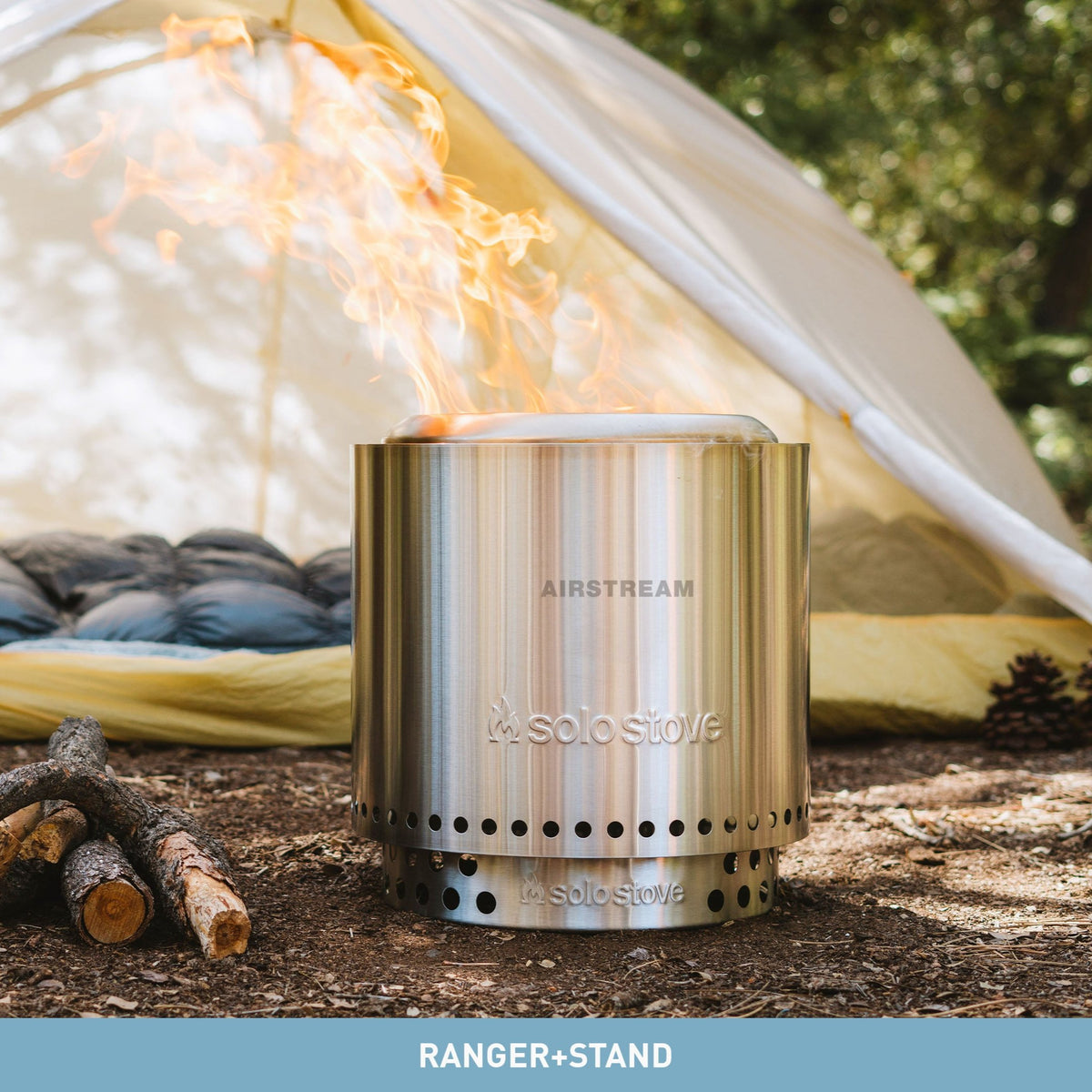 An Airstream Ranger Fire Pit by Solo Stove burns outside near a tent in a wooded campsite, with small logs stacked nearby. Thanks to its 360° Airflow design, the portable stainless steel fire pit delivers an efficient, low-smoke burn. The tent is open, revealing dark bedding inside.