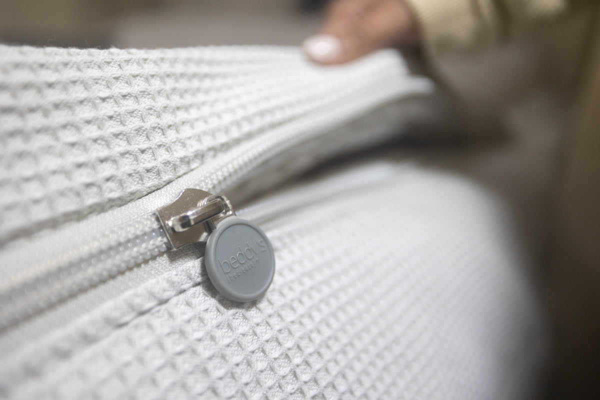 A close-up of a textured white fabric with a zipper. A hand is holding the fabric, and the zipper pull is labeled bed sheets..