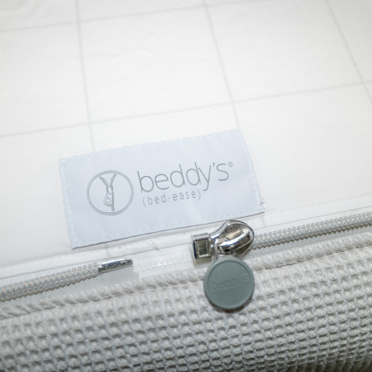 Close-up of a white Airstream Custom Fit Beddy’s for Globetrotter Travel Trailers bedding label and zipper pull on a textured quilt. The label reads beddys (bed-ease), and there is a round zipper pull featuring the Beddys logo—showcasing the all-in-one zippered bedding designed for convenience, ideal for compact spaces like those in Airstream travel trailers.