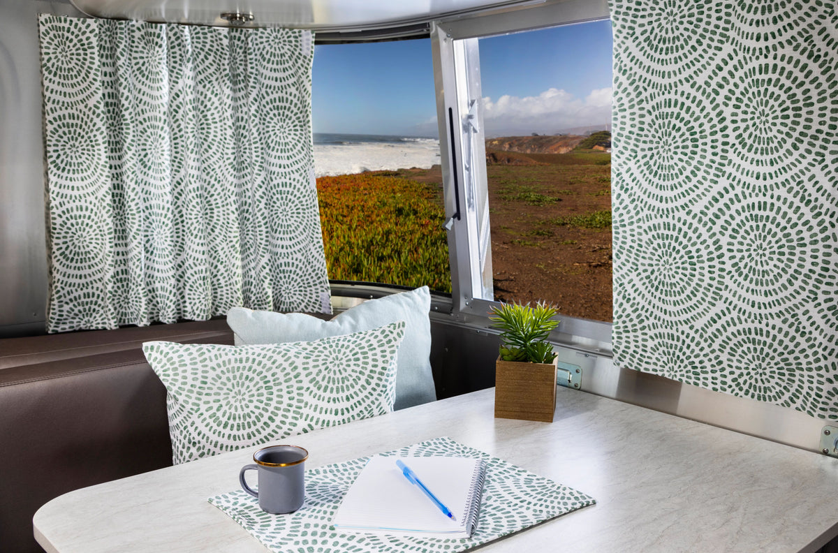 A table with a mug, notepad, pen, and plant sits inside an RV with patterned cushions and Airstream Printed Custom Curtains for Sport Travel Trailers. Through the window, there’s a stunning view of the coast with waves, cliffs, and blue sky.
