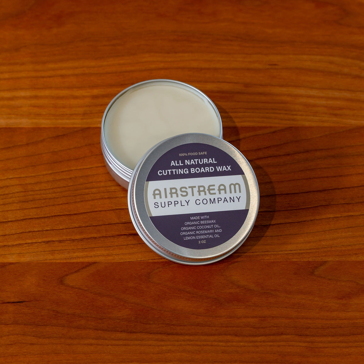 An open round tin labeled Airstream Supply Company All-Natural Cutting Board Wax rests on a wooden surface, displaying the wax inside. Perfect for maintaining your Airstream Custom Sink Cutting Boards for Basecamp, the lid features that it is food safe and made with organic oils—making it an ideal companion to your collection of Airstream Basecamp accessories.