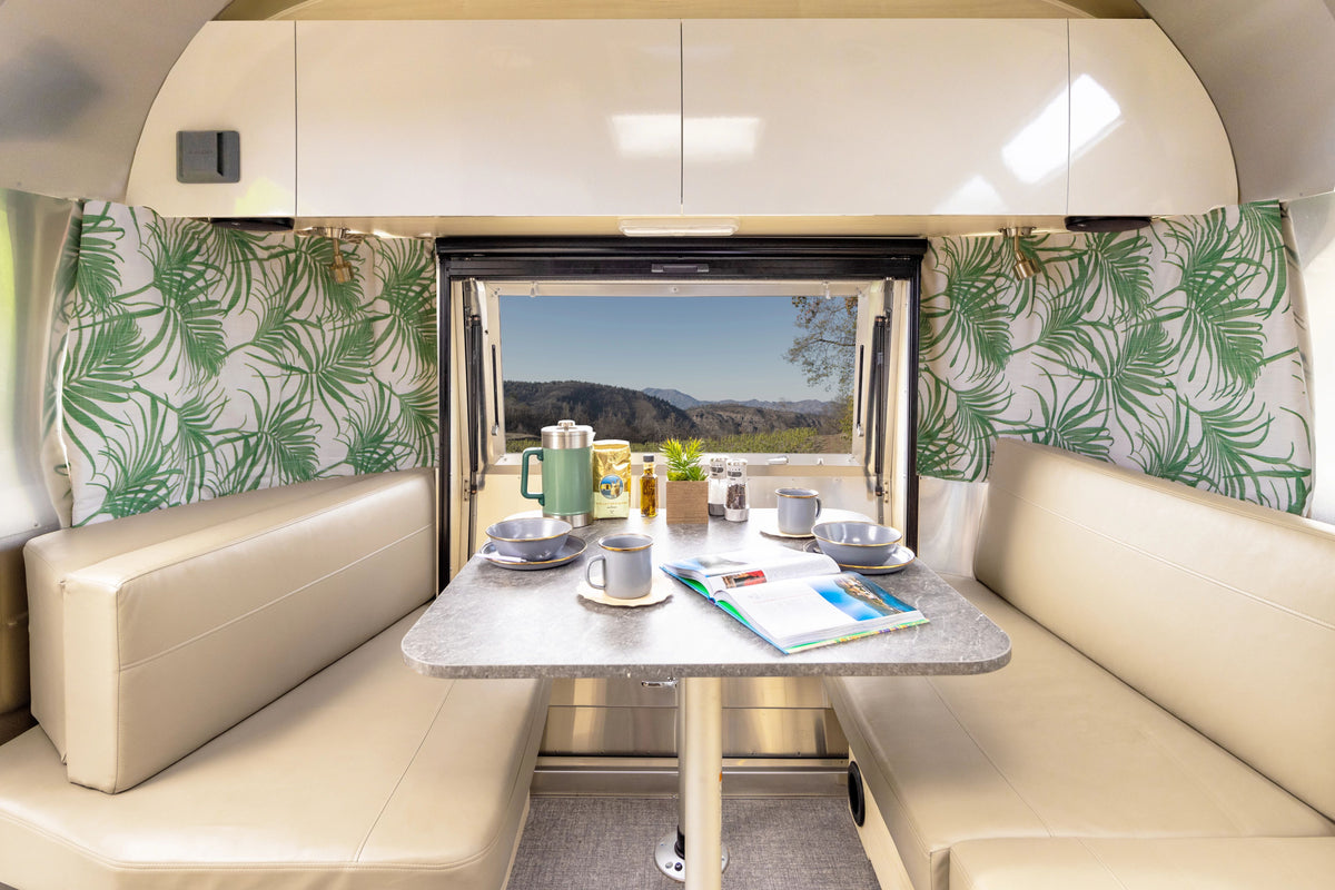 A cozy RV dining area with beige seats, a set table with coffee and dishes, Airstream Printed Custom Curtains for International Signature Travel Trailers in a leafy green pattern, and a window framing a scenic mountain view. A book and magazine lie open on the table.
