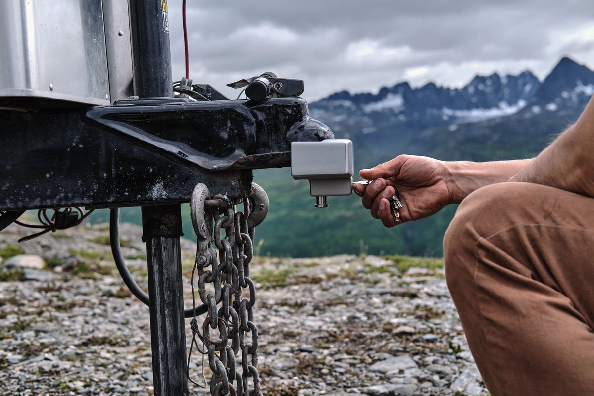 A person kneels outdoors, securing an Airstream travel trailer with the Airstream Anti-Theft Hitch and Trailer Lock Kit, ensuring protection against theft, with mountains and a cloudy sky in the background.