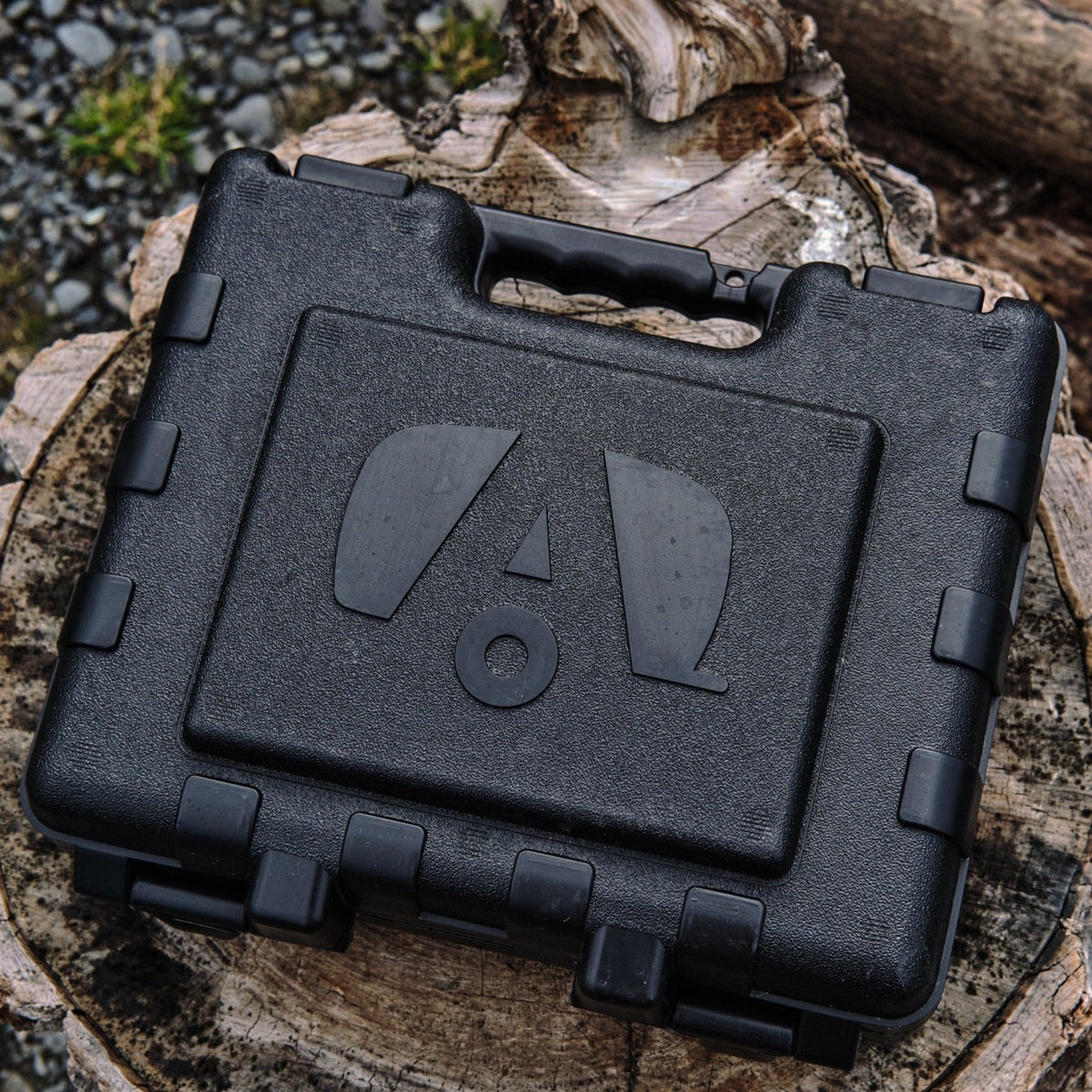 A black hard plastic carrying case with a handle and embossed logo, designed to hold the Airstream Anti-Theft Hitch and Trailer Lock Kit, sits on a cut tree stump outdoors, surrounded by logs and gravel—a reliable solution for securing your Airstream travel trailer against theft.