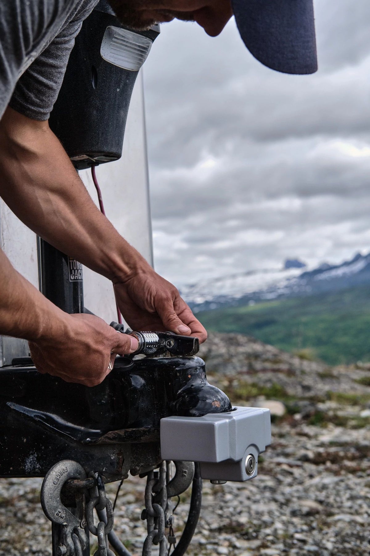 A person wearing a cap tightens a bolt on a trailer hitch outdoors, with mountains and a cloudy sky in the background, securing their setup with an Airstream Anti-Theft Hitch and Trailer Lock Kit for enhanced Airstream security.