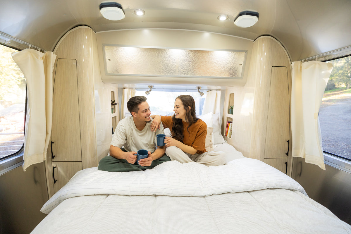 A couple sits on a bed inside a camper van, smiling and holding mugs. They face each other, surrounded by light wood cabinets and large windows with cream curtains, creating a cozy and bright atmosphere.