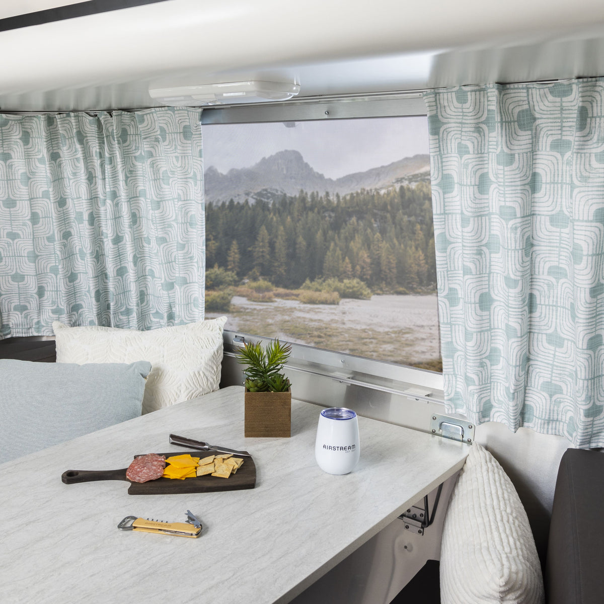 A table inside a camper displays a cheese and meat tray, a small plant, and a white canister. The window reveals trees and mountains perfectly framed by Airstream Printed Custom Curtains for Pottery Barn Travel Trailers in an elegant blue-and-white pattern.