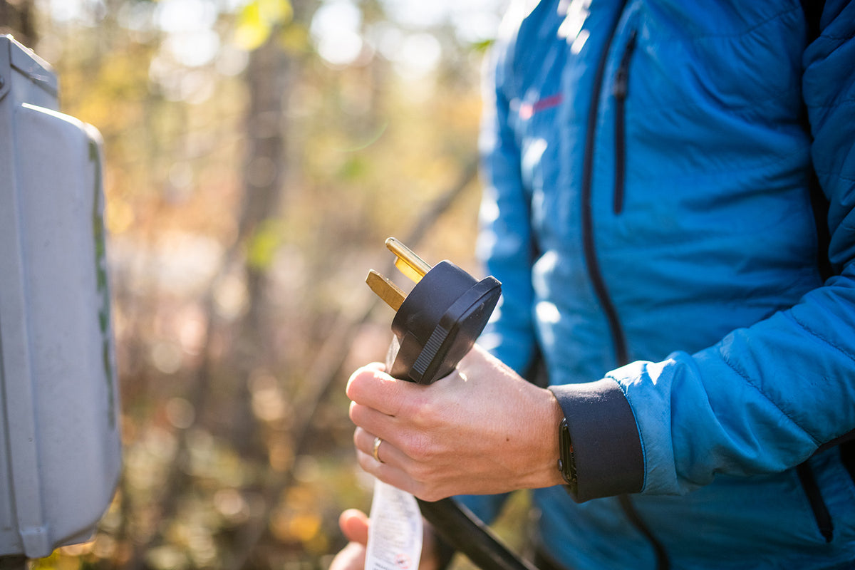 A person in a blue jacket holds an electrical plug near a power outlet outdoors, preparing to connect it.