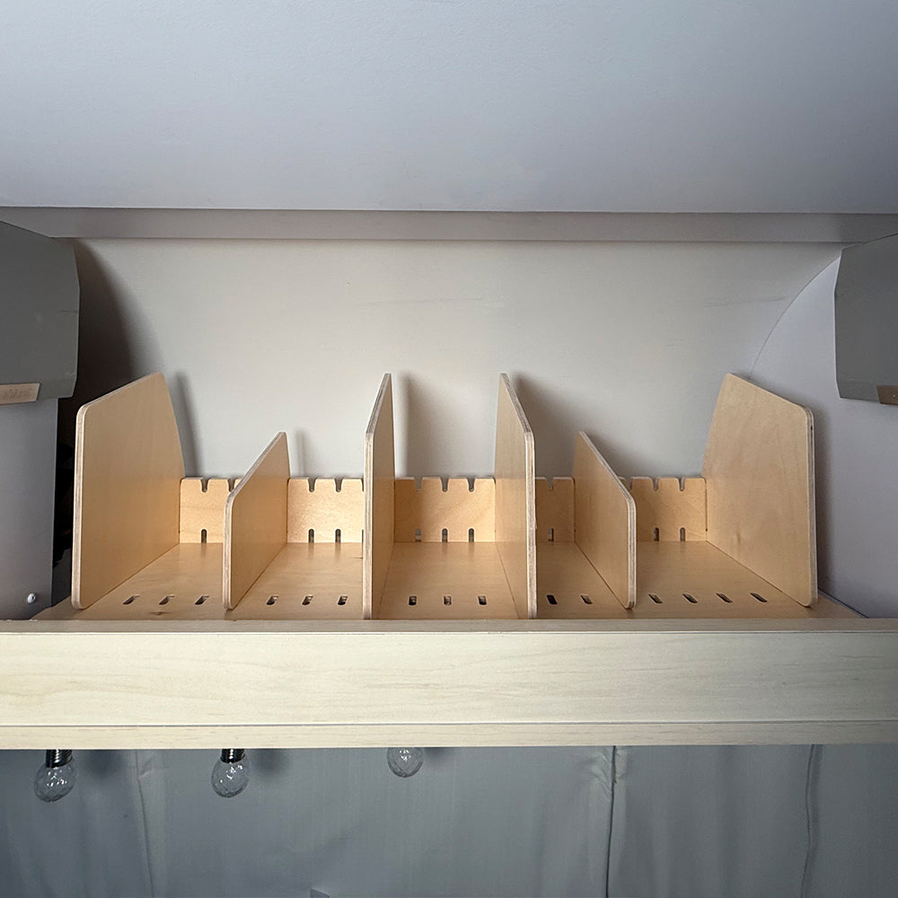 Airstream Roof Locker Organization by Airworthy Equipment Company, featuring six adjustable vertical dividers, mounted against a white wall. Inspired by classic Airstream roof locker design, the shelf is empty, with light bulbs hanging beneath its edge.