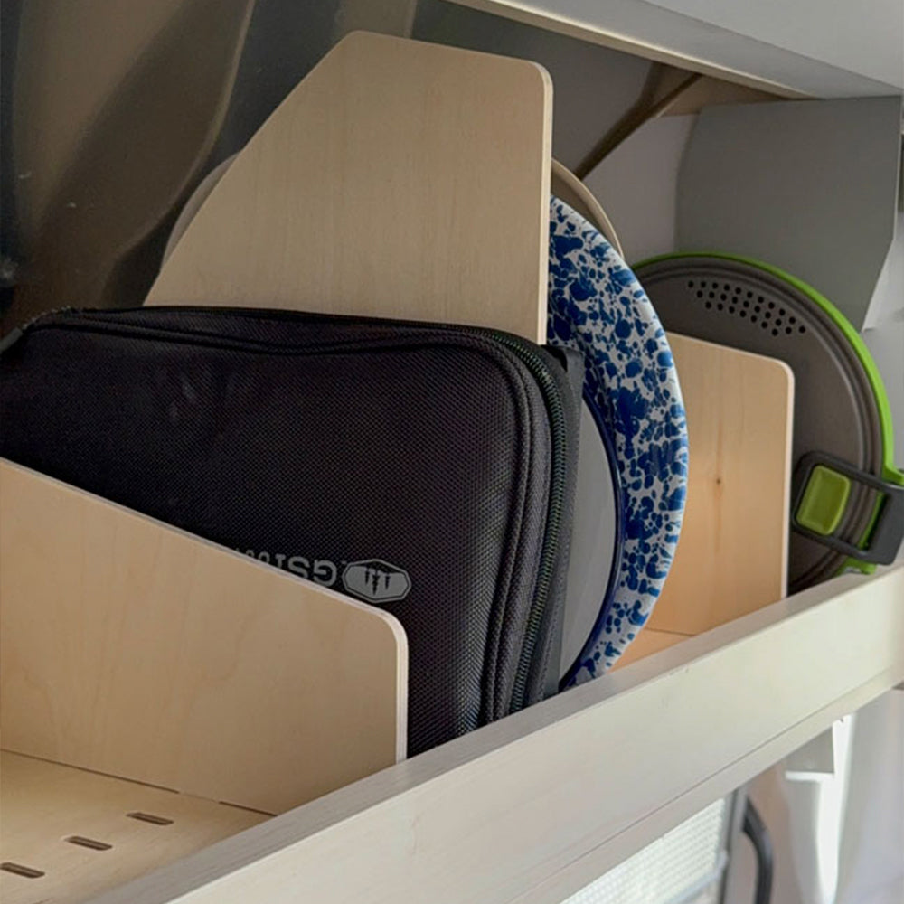The Airstream Touring Coach Roof Locker Organization by Airworthy Equipment Company features wooden dividers that neatly arrange dishes and a black zippered bag on the shelf, maximizing storage efficiency. Plates with blue floral and green designs are stored upright in individual sections, creating an organized space.