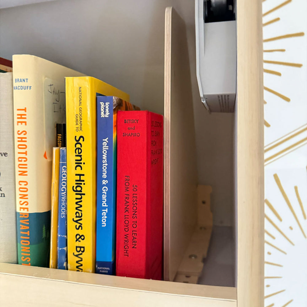 Books are neatly arranged on a wooden shelf, separated by a vertical divider, forming the Airstream Touring Coach Roof Locker Organization by Airworthy Equipment Company. The visible book titles include The Shotgun Conservationist and Yellowstone & Grand Teton, with part of a patterned wall seen on the right.