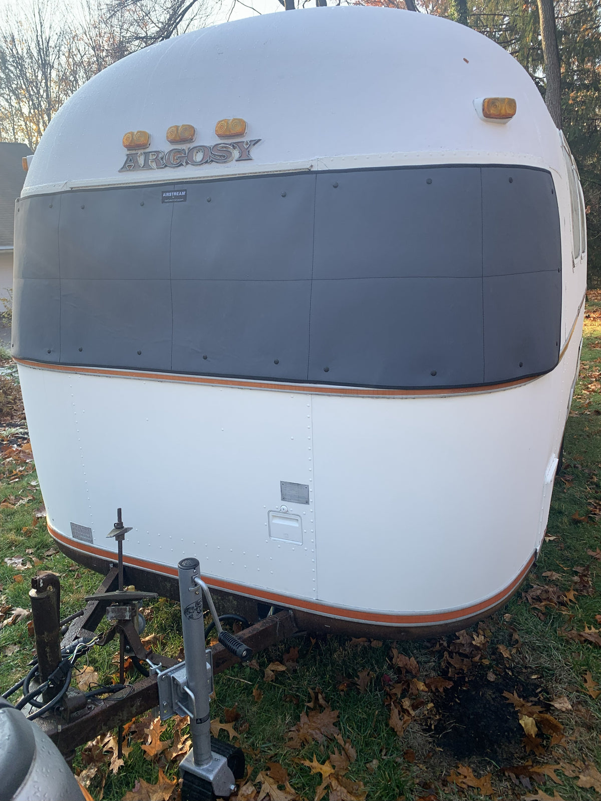 A white Argosy travel trailer parked on the grass, with an Airstream Padded Window Protector for Argosy—a black panoramic cover—draped over its front windows, autumn leaves scattered around it, making a perfect showcase of vintage travel trailer accessories.