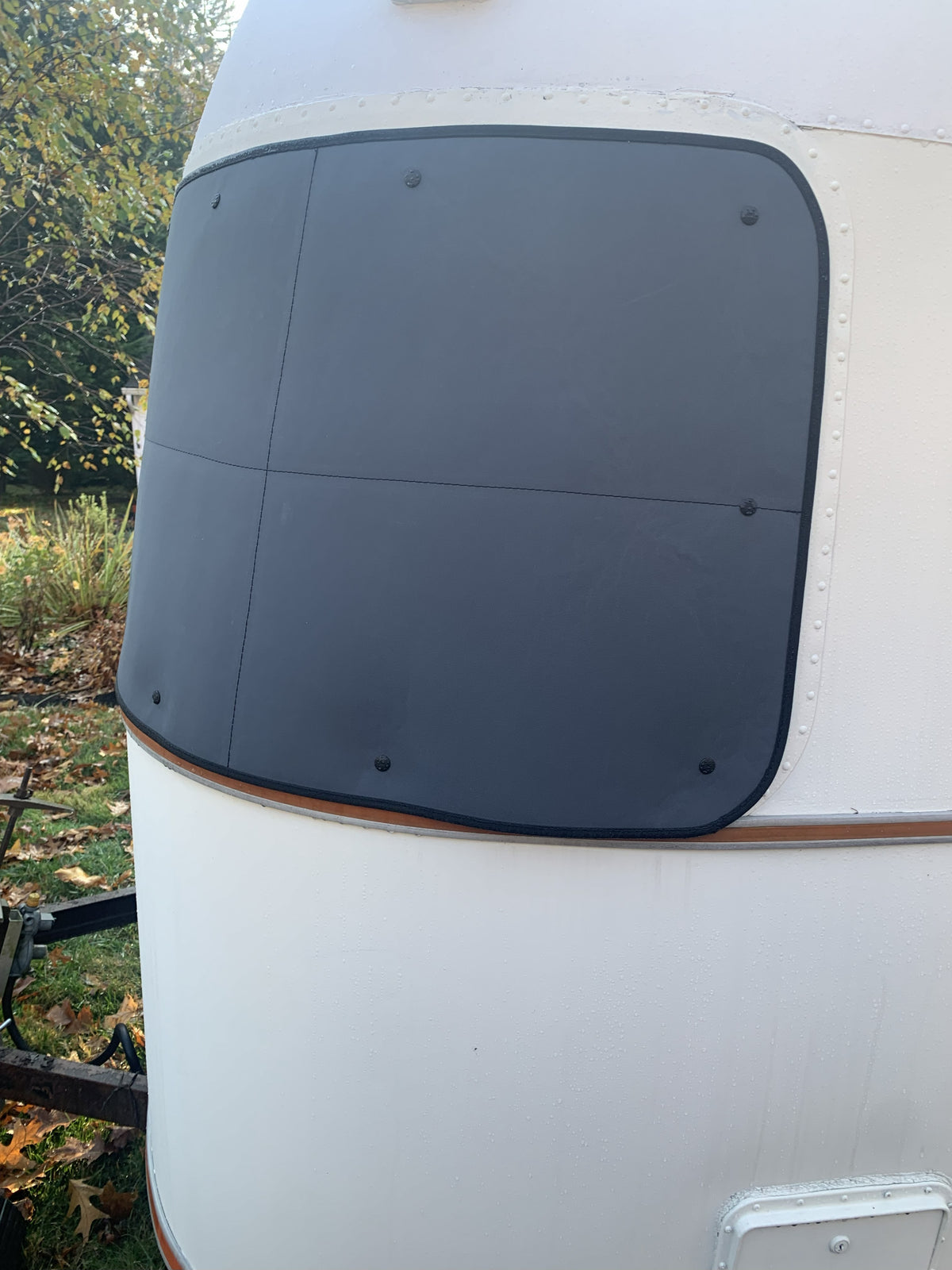 Close-up of the Airstream Padded Window Protector for Argosy, a matte black cover securely attached to the front window of a white travel trailer. As one of the essential vintage travel trailer accessories, it enhances both style and protection. In the background, trees and fallen leaves can be seen.
