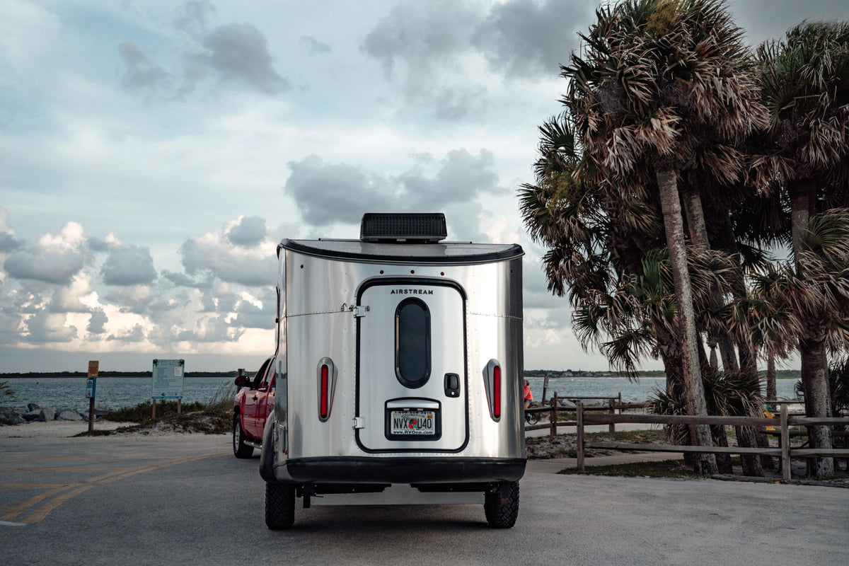 A silver Airstream trailer, equipped with the Airstream Main Door and Cargo Door Pre-Cut Key for Basecamp (WD Series), is hitched to a red truck and parked near palm trees by the water under a cloudy sky at a coastal location.