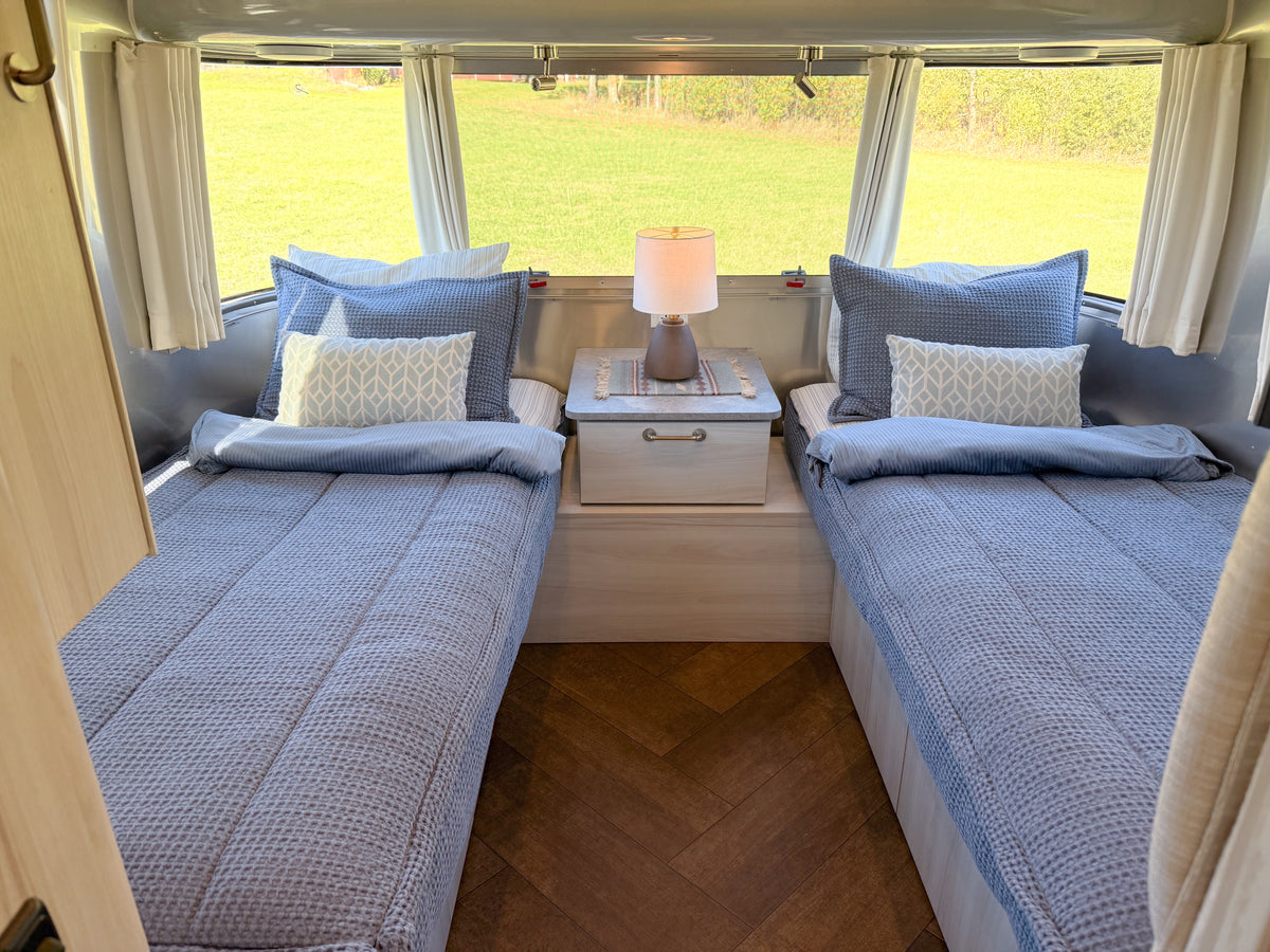 Two twin beds in a travel trailer outfitted with blue textured Airstream Custom Fit Dual Comfort Beddy’s for International Travel Trailers and decorative pillows, separated by a nightstand with a lamp.