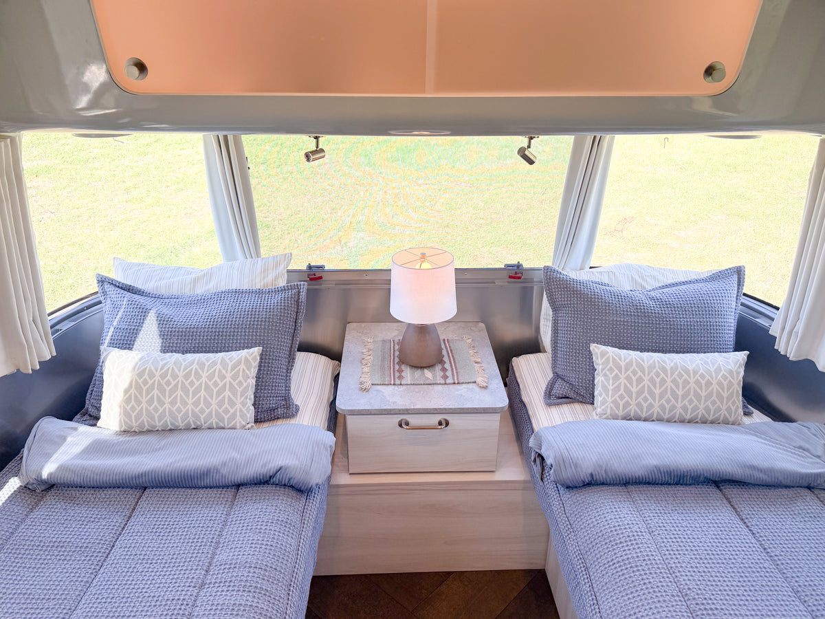 Two twin beds in a travel trailer outfitted with Airstream Custom Fit Dual Comfort Beddy’s for International Travel Trailers, each made with blue waffle-weave bedding and a patterned accent pillow, separated by a nightstand with a lamp.
