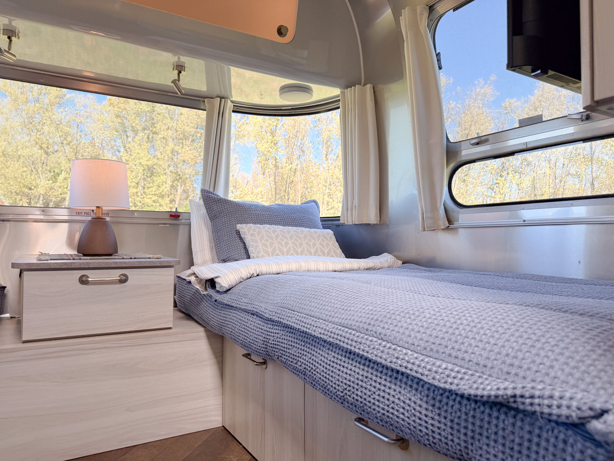 A twin bed in a travel trailer featuring Airstream Custom Fit Dual Comfort Beddy’s for Caravel Travel Trailers with a blue textured duvet, coordinating shams, and a bedside lamp.