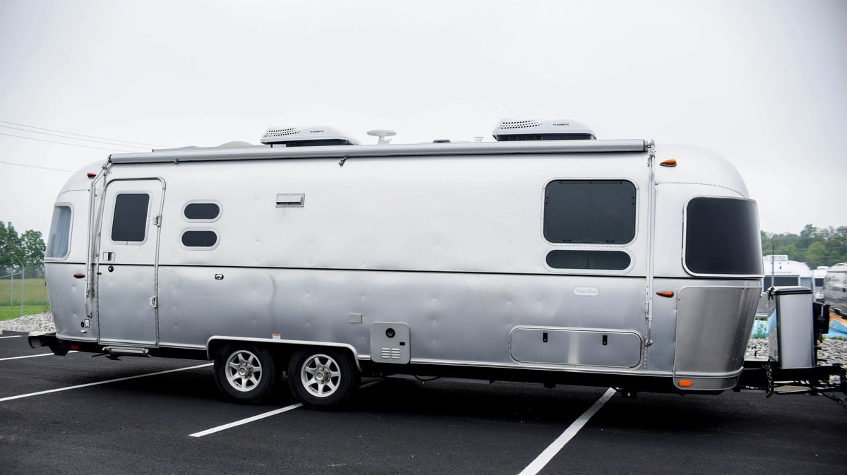 An Airstream travel trailer, accented with sleek aluminum Premium Replacement Belt Trim from Airstream, is parked in a blacktop lot with white parking lines under a cloudy sky. Trees and other trailers can be seen in the background.