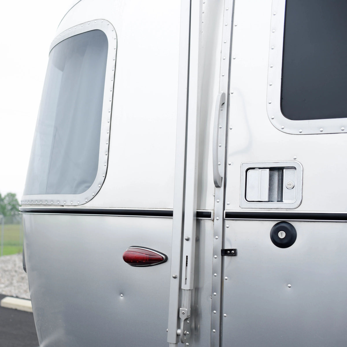 Close-up of a silver Airstream trailer door and window, featuring metal rivets, a handle, a lock, a small red tail light, and the sleek Airstream Premium Replacement Belt Trim for Travel Trailers as an exterior accessory.