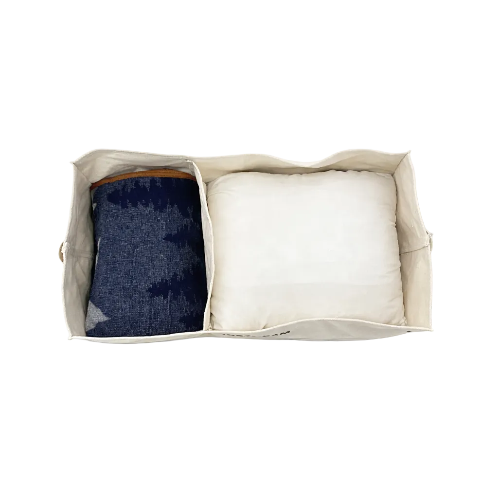 The Airstream x Steele Canvas Twin Bed Storage Bin, shown from above with a folded dark blue patterned blanket on the left and a white pillow on the right, is perfect for organizing essentials in your twin bed floor cabinet.