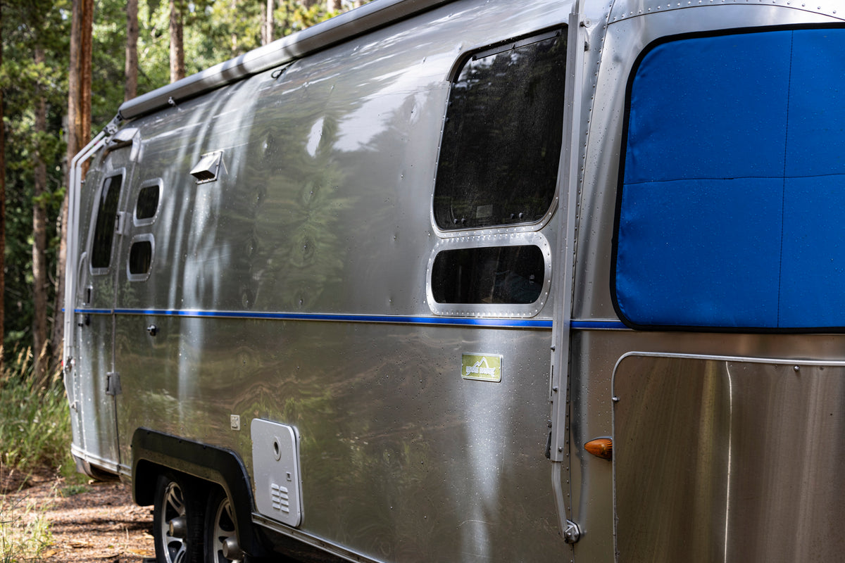 A shiny silver camper trailer with blue window coverings is parked in a wooded area, surrounded by tall green trees and grass. The Airstream Premium Replacement Belt Trim for Travel Trailers gleams in the sunlight, accentuating the classic design of this vintage retreat.