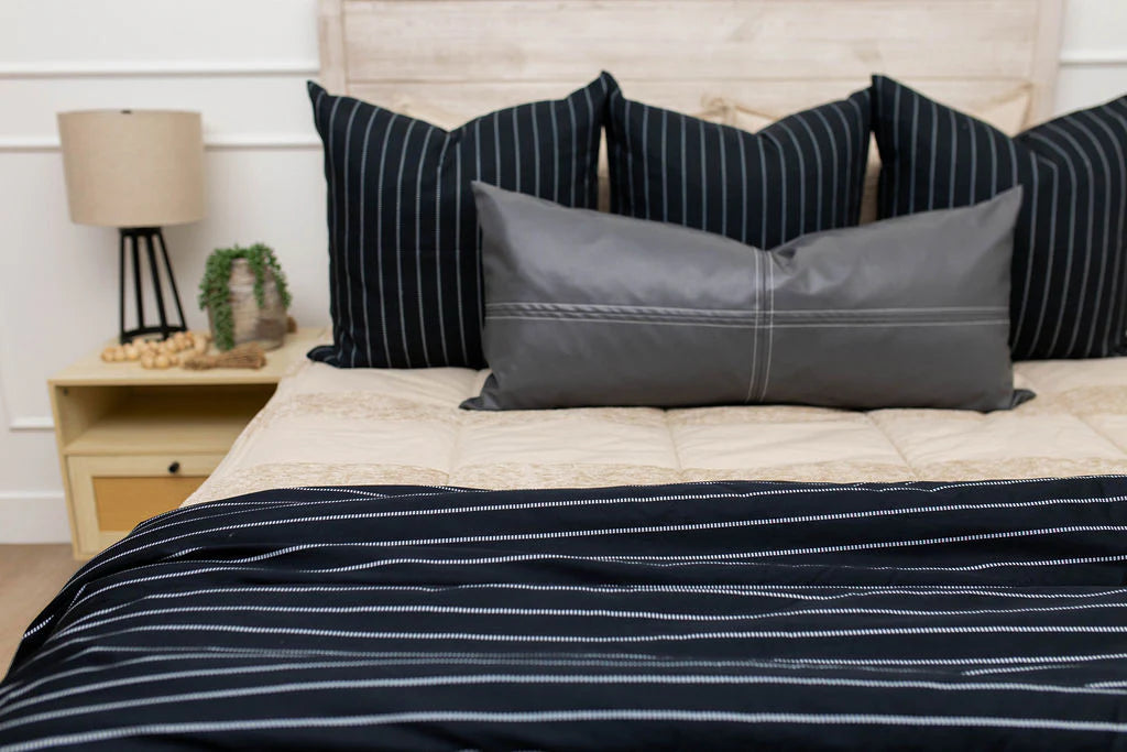 A neatly made bed features black and gray striped pillows, a gray bolster, beige bedding, and the Bryant Luxe Blanket by Beddy’s draped across it, showcasing its modern black striped design. To the left of the bed is a nightstand topped with a lamp and a small potted plant.