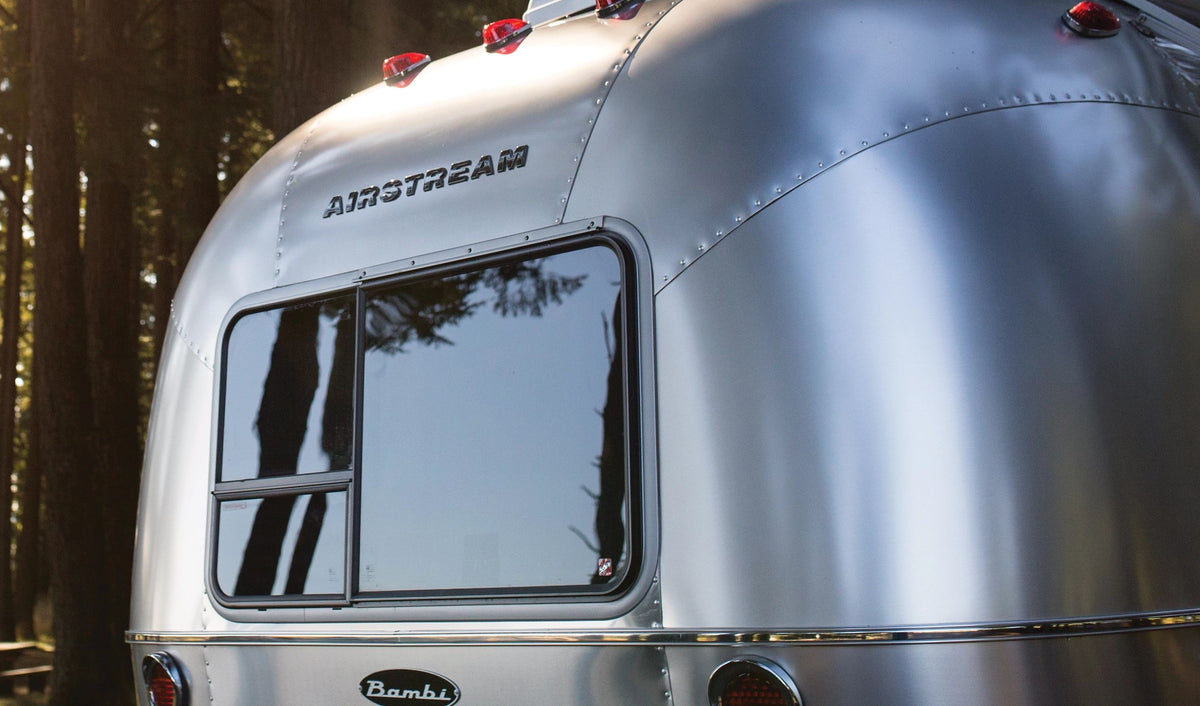 The rear of a shiny silver Airstream trailer with a large window and the word AIRSTREAM above it, parked outdoors among tall trees.
