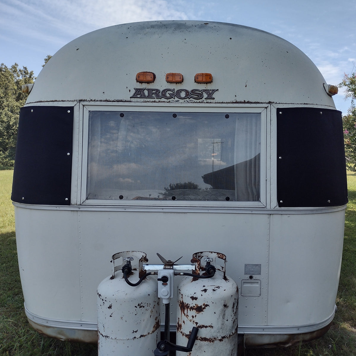Rear view of a white Argosy travel trailer with two rusted propane tanks mounted below a large window fitted with an Airstream Padded Window Protector for Argosy, set on grass with trees and sky in the background.