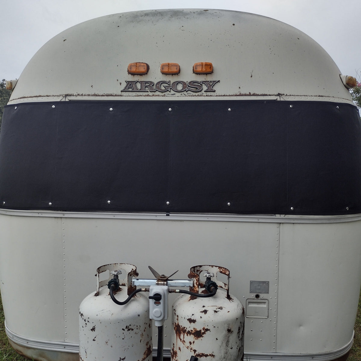 The photo shows the back of a white vintage Argosy trailer with two rusted propane tanks mounted at the bottom, featuring an Airstream Padded Window Protector for Argosy as one of its classic vintage travel trailer accessories.