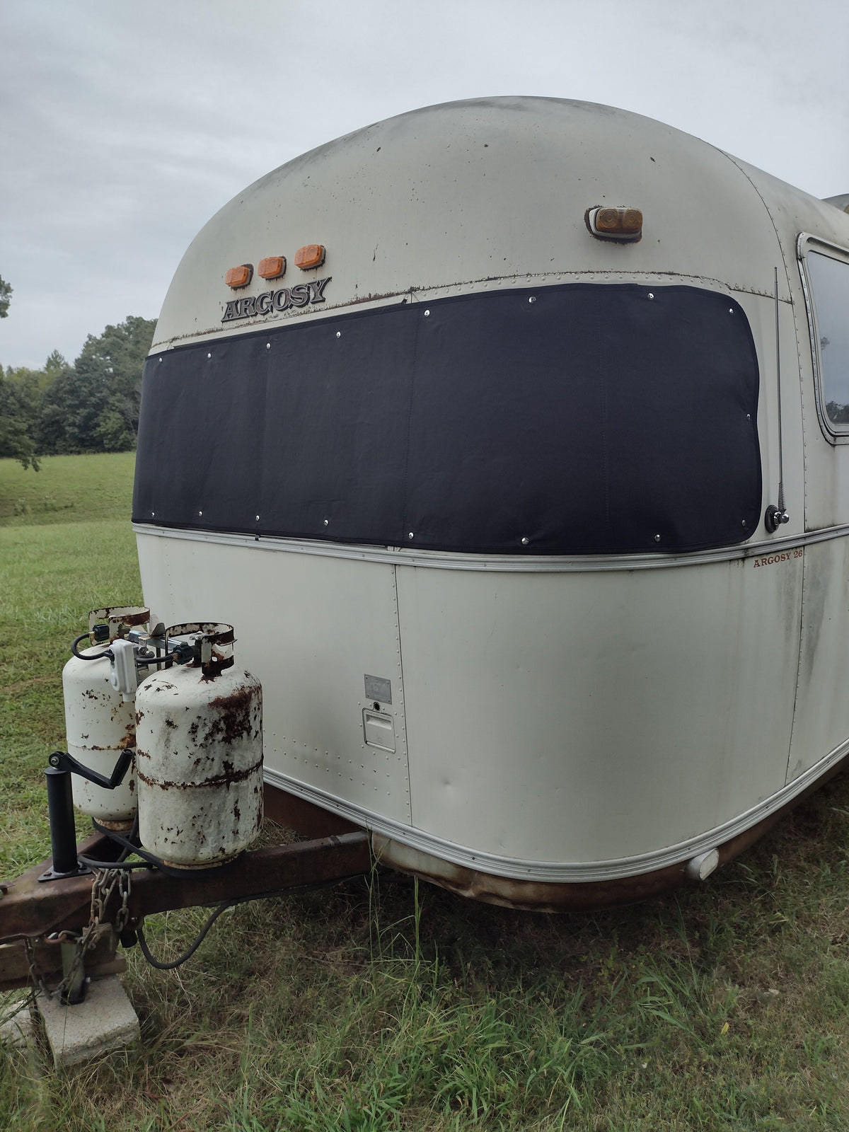 A vintage Argosy travel trailer with propane tanks attached at the front is parked on grass, surrounded by trees in the background. Its large panoramic windows are shielded by an Airstream Padded Window Protector for Argosy—one of the essential accessories for vintage travel trailers.