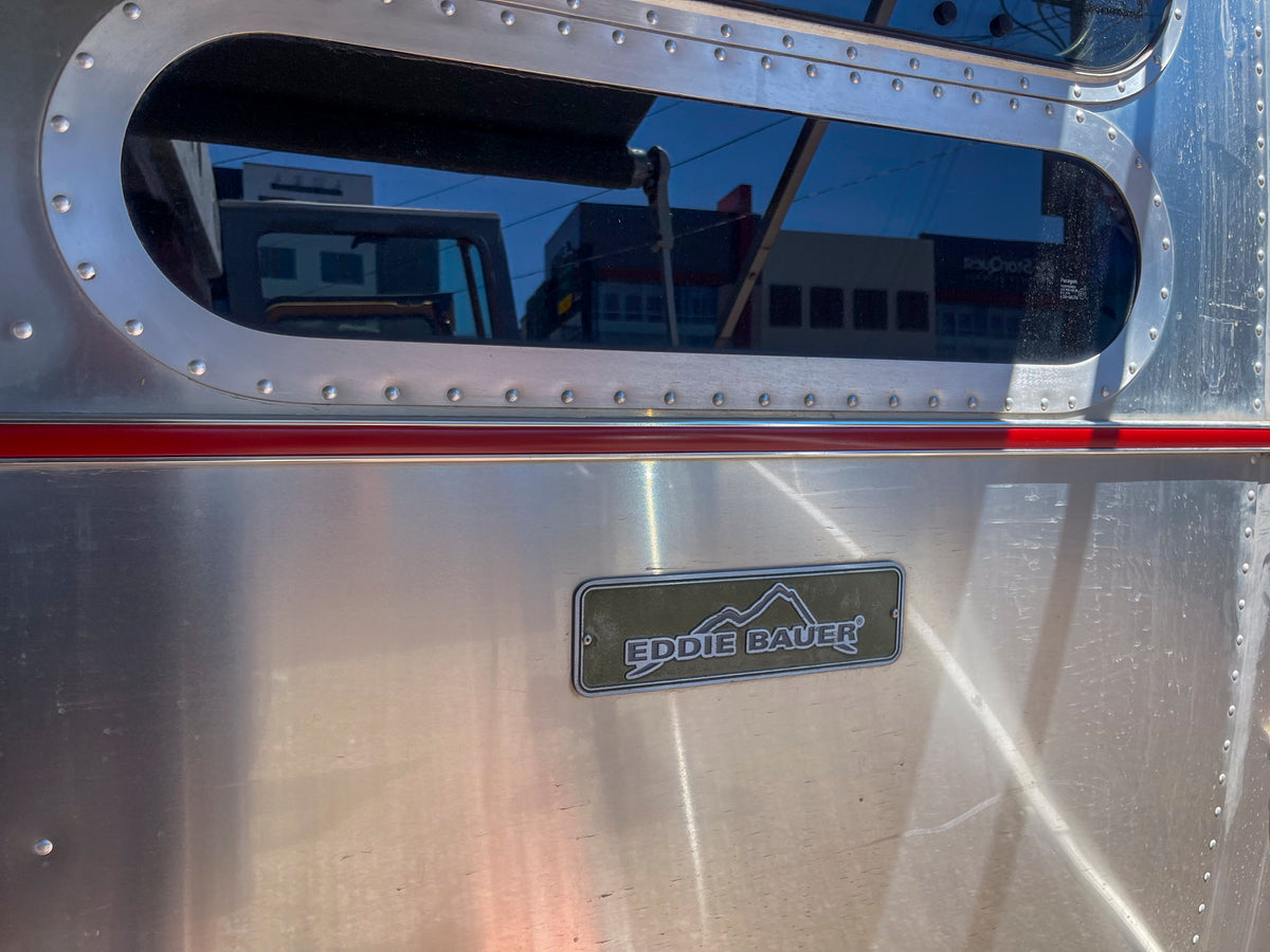 Close-up of a shiny metallic surface featuring the Airstream Premium Replacement Belt Trim for Travel Trailers with a red stripe, a window reflection, and a rectangular badge reading EDDIE BAUER with mountains above the text—an ideal exterior accessory for those seeking authentic Airstream style.