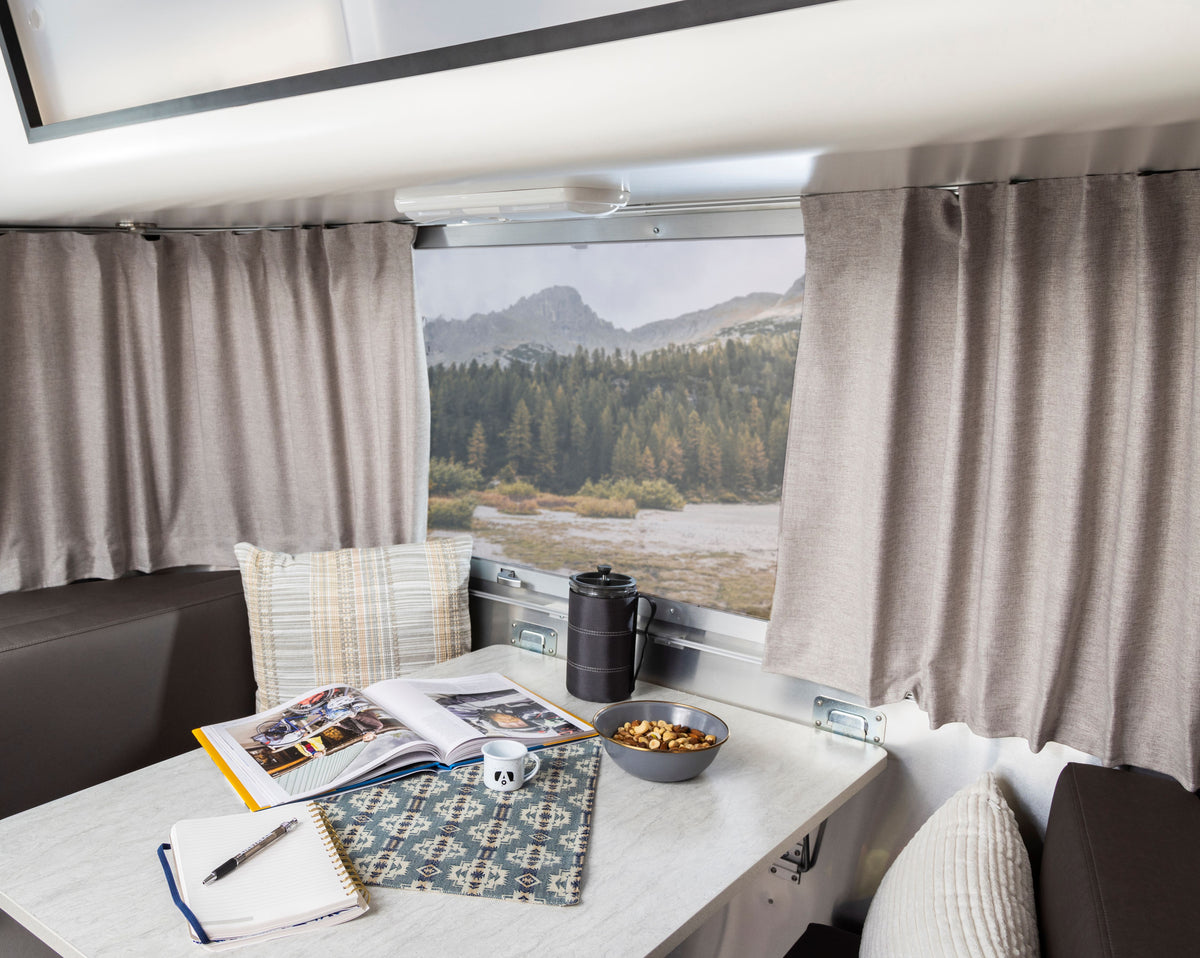 A cozy RV dining area features a marble table adorned with books, a coffee pot, a bowl of snacks, and a pillow. The window offers views of mountains and forest, beautifully framed by Airstream Solid Custom Curtains for Bambi Travel Trailers in soft gray.