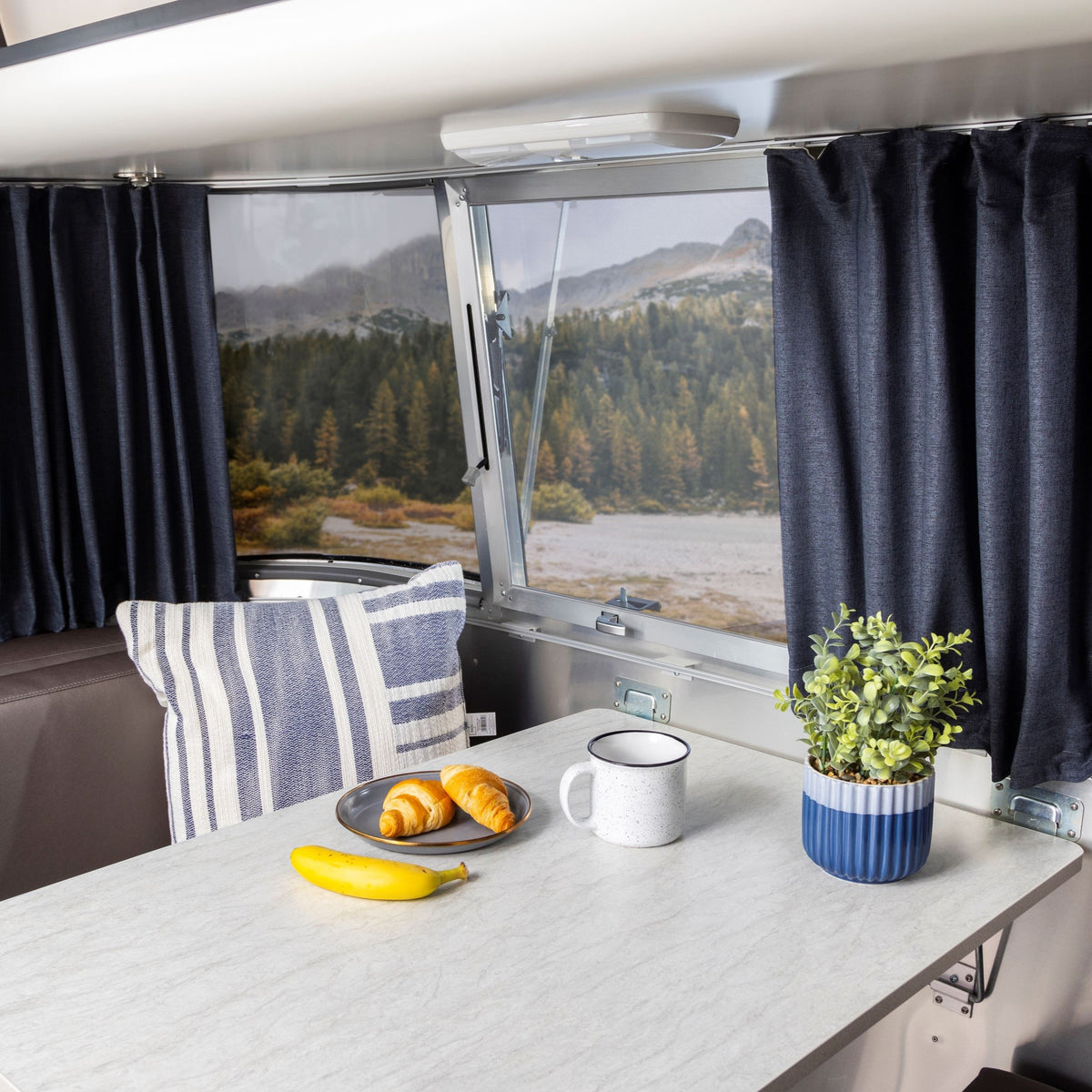 A table in an RV holds a banana, two croissants, and a mug. A potted plant and a striped pillow add charm, while mountains and trees can be seen through the window. The window is framed by Airstream Solid Custom Curtains for International Travel Trailers, which bring warmth and style to the inviting space.