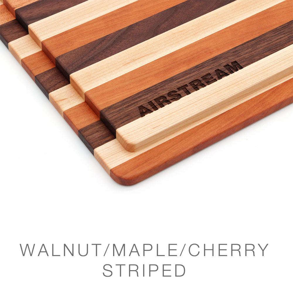 A close-up of striped Airstream Custom Sink Cutting Boards for Basecamp, crafted from walnut, maple, and cherry wood. One board features “AIRSTREAM” engraved on a maple stripe—perfect for use as a sink cutting board in your travel trailer. Text below reads WALNUT/MAPLE/CHERRY STRIPED.