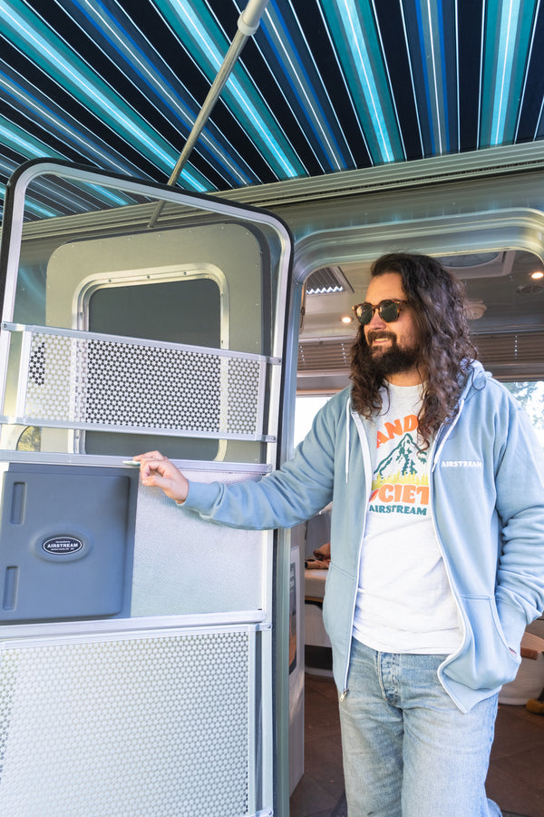 A man with long hair and sunglasses smiles while standing at the open door of an Airstream RV under a striped blue awning, wearing an Airstream Wanderer Society Long Sleeve T-Shirt.