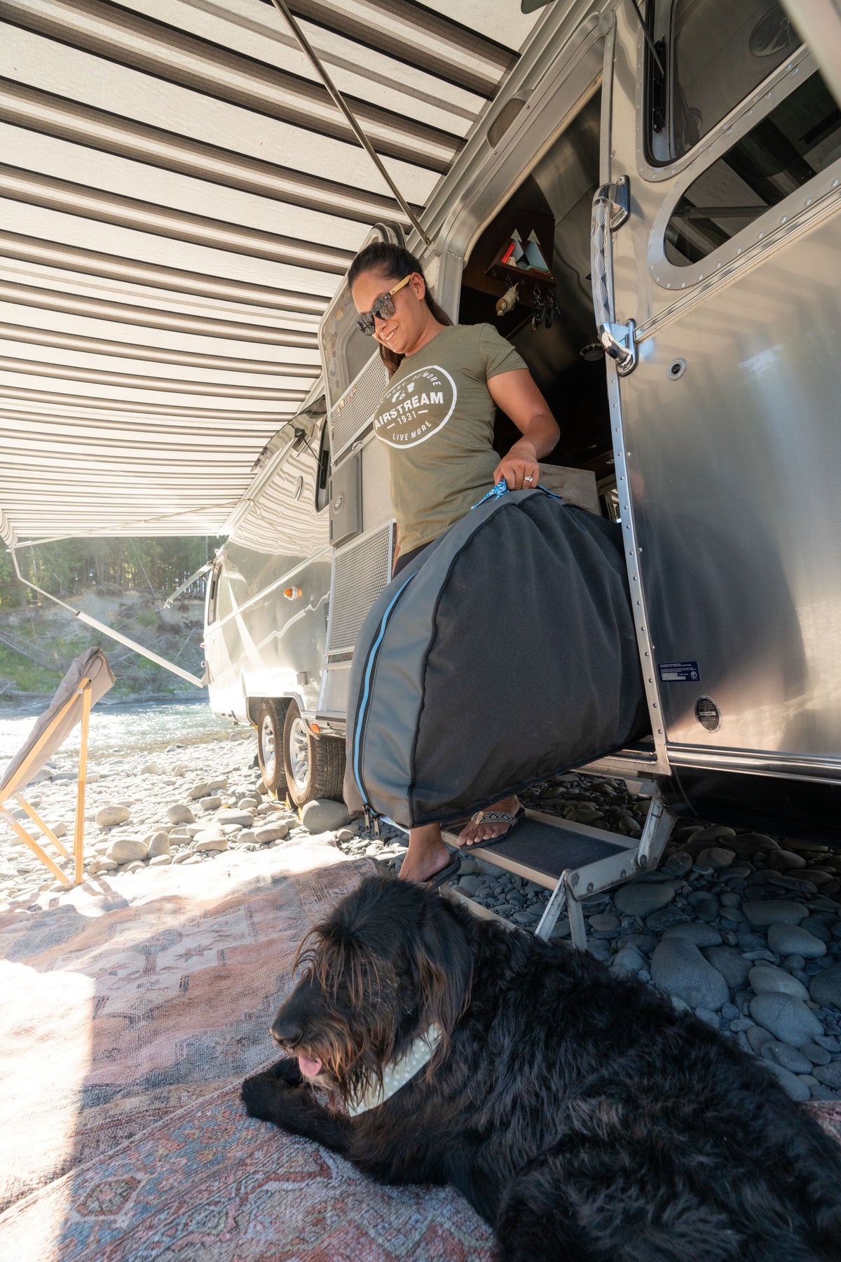 A woman steps out of an Airstream trailer holding a large bag, while a large black dog relaxes on the Airstream Travel Pet Bed outside on the rocky ground.