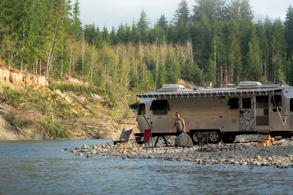 A person stands by a silver RV parked on a rocky riverbank, surrounded by forest. Two chairs face the water, and an Airstream Travel Pet Bed sits nearby, providing comfort for their furry companion. The scene exudes a peaceful, outdoor camping vibe.