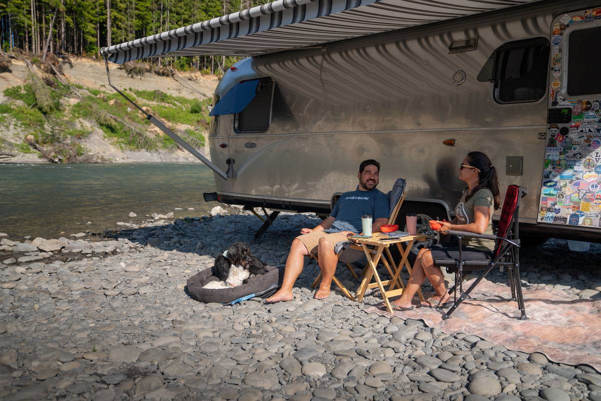 A man and woman relax in chairs beside their camper van on a rocky riverside, dining at a small table. Nearby, their black and white dog lounges comfortably on an Airstream Travel Pet Bed. Trees and water form a tranquil backdrop.