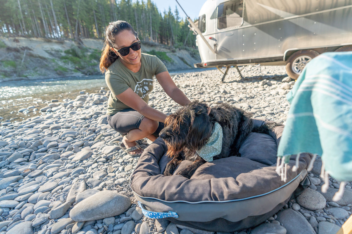 Wearing sunglasses, a woman smiles at a black dog lounging in the Airstream Travel Pet Bed on a rocky riverbank, with a trailer and trees behind them—capturing the spirit of an outdoor camping adventure.