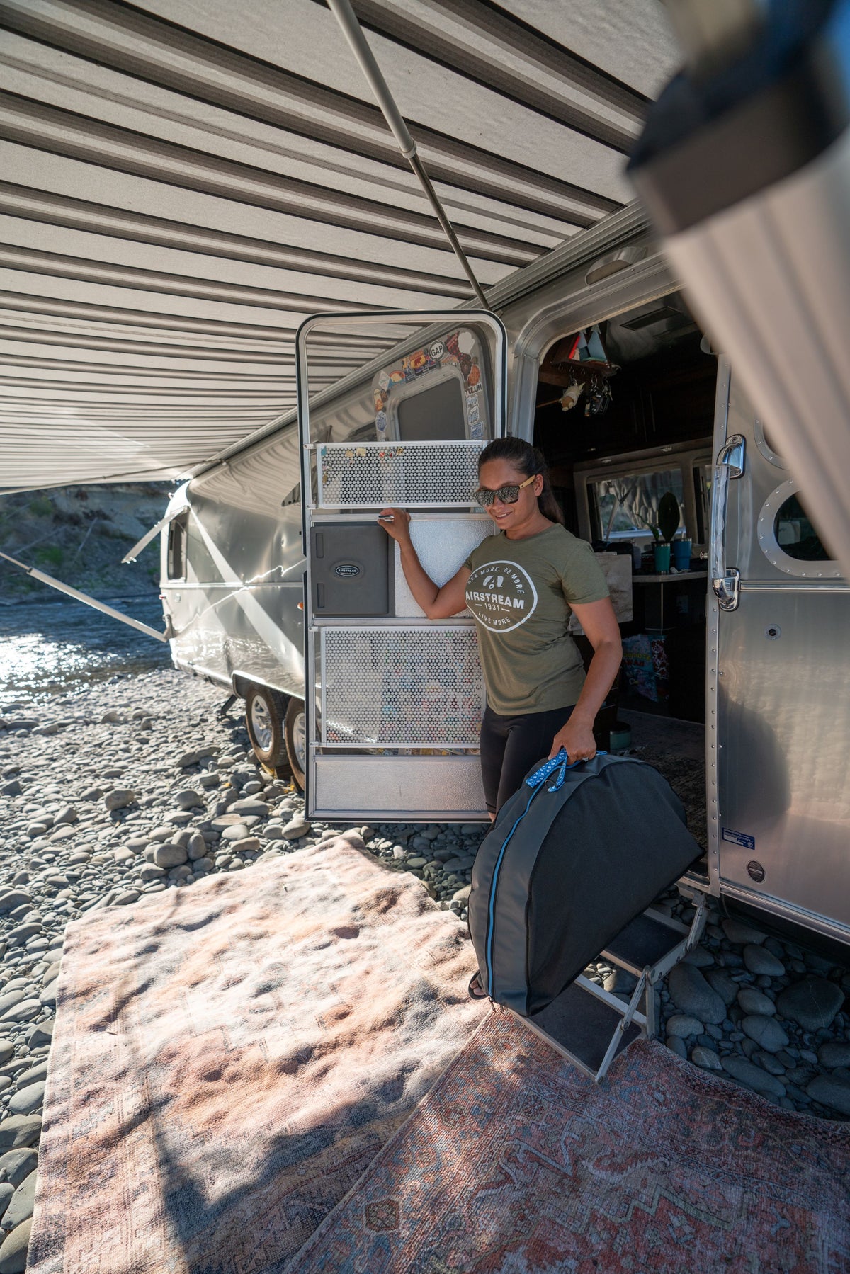 A woman wearing sunglasses stands at the door of a silver camper, holding a folded chair under an awning, with a patterned rug and an Airstream Travel Pet Bed on the rocky ground outside by a river.