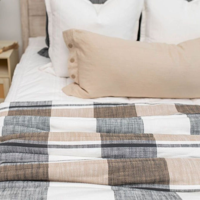 A neatly made bed with white and beige pillows and a beige lumbar pillow features a plaid comforter in shades of white, beige, and gray. Draped across the foot is the Erin Luxe Blanket by Beddy’s, adding an extra layer of softness. A light wood nightstand is partially visible on the side.