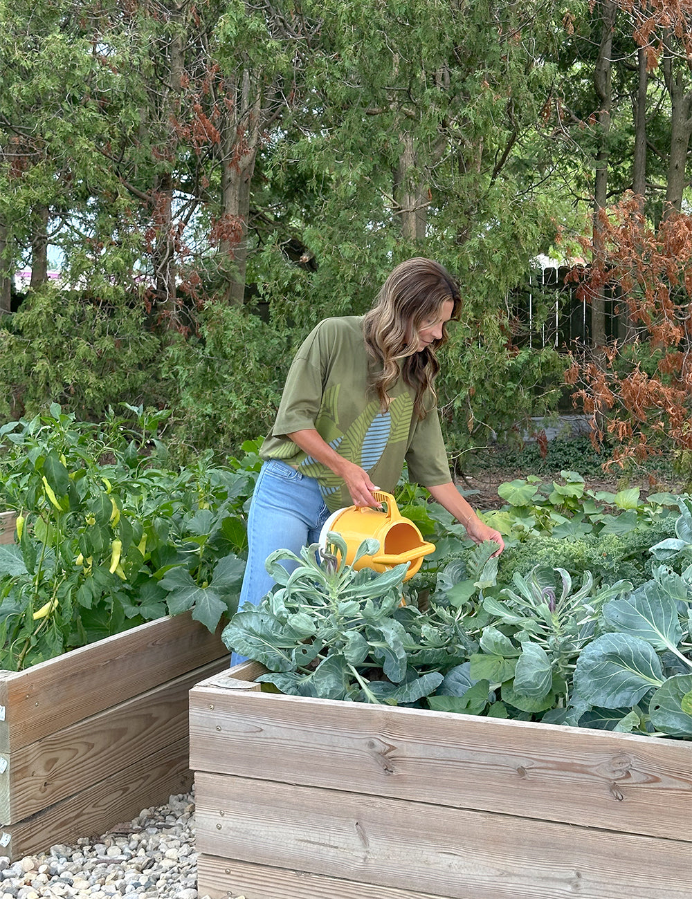 Wearing the Frank Lloyd Wright x Airstream Oversized T-Shirt and a blue skirt, a woman waters leafy vegetables in a raised garden bed with a yellow watering can. She stands outdoors among lush green plants and trees.