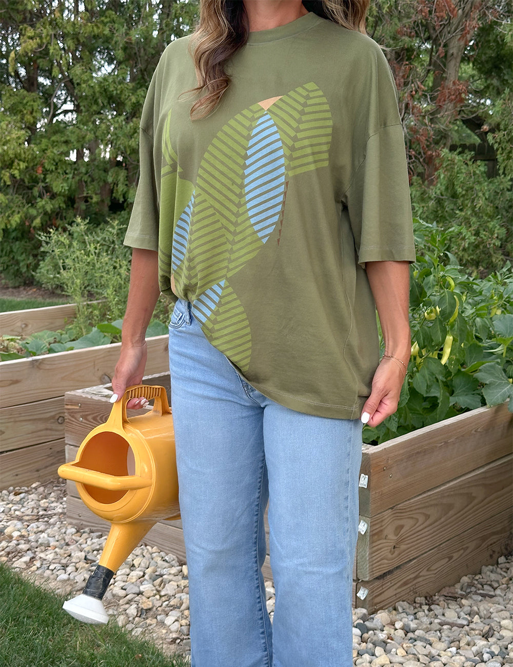 A person stands in a garden holding a yellow watering can, wearing the Frank Lloyd Wright x Airstream Oversized T-Shirt with a green graphic design and blue jeans. Raised garden beds and green plants are visible in the background.