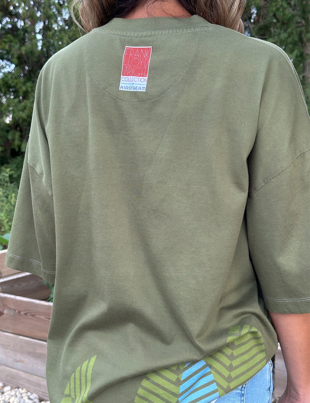 A person is wearing the Frank Lloyd Wright x Airstream Oversized T-Shirt in olive green, which features a red and white label on the upper back. Inspired by architectural graphic t-shirt designs, the shirt displays blue and green leaf patterns near the bottom. The background includes greenery and wooden planter boxes.
