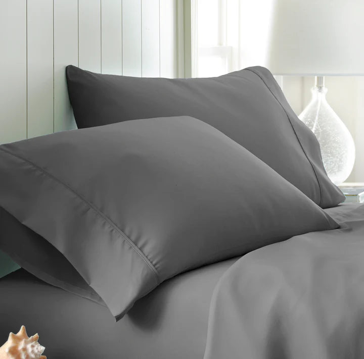 Two gray Cotton Percale 200 Thread Count Pillowcases rest on a neatly made bed with matching cotton percale sheets, offering a crisp and comfortable feel. A white paneled wall and a glass lamp are in the background, with a seashell decor piece visible in the lower left corner.