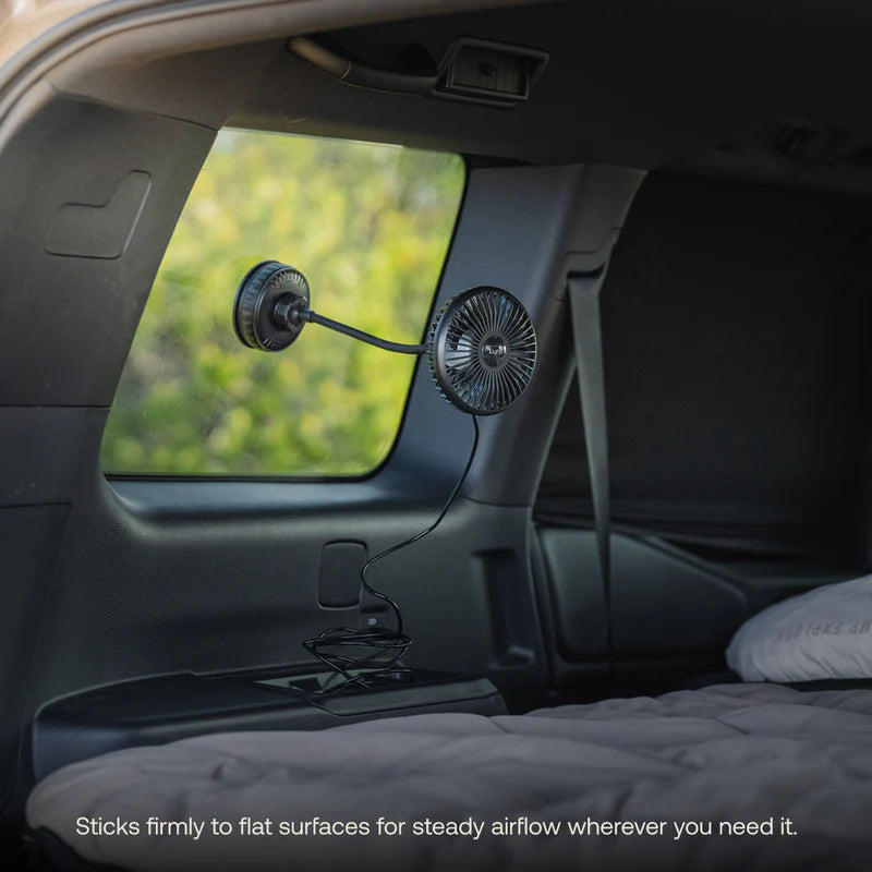 A USB Powered Camping Fan by Luno, attached to a van window with a suction mount and powered via USB-C, is positioned near the bed inside the vehicle. Ideal for car camping or van life, this setup provides comfort while allowing you to enjoy views of trees outside through the glass.