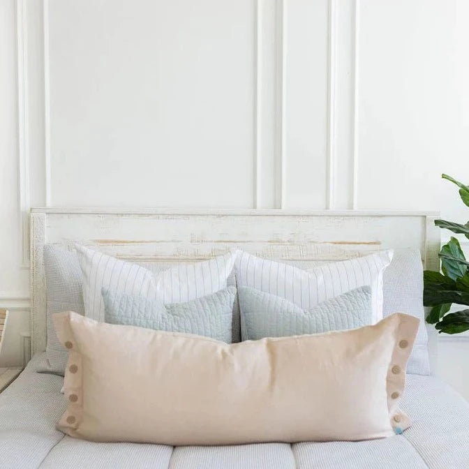 A neatly made bed features two blue and white decorative throw pillow covers, with the Harper Medium Pillow Cover by Beddy’s—a large beige accent pillow cover—arranged in front, set against a white wooden headboard and a white paneled wall, with a green plant placed on the right side.