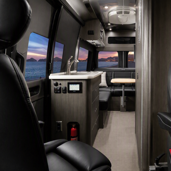 The interior of a luxury motorhome or van, featuring two black leather passenger seats on either side of a narrow aisle, a kitchenette with a sink and countertop on the left, and a dining area in the back with a table and bench. The walls are finished in a wood grain pattern, and the floor is a light beige carpet.