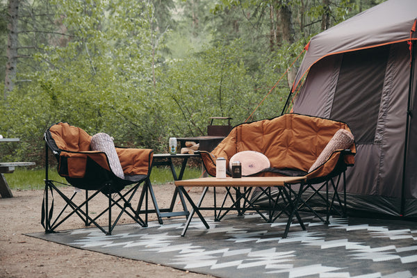 Outdoor camping setup with two tan and black folding camp chairs, a matching folding coffee table, a small side table, and a gray tent in a forest clearing.